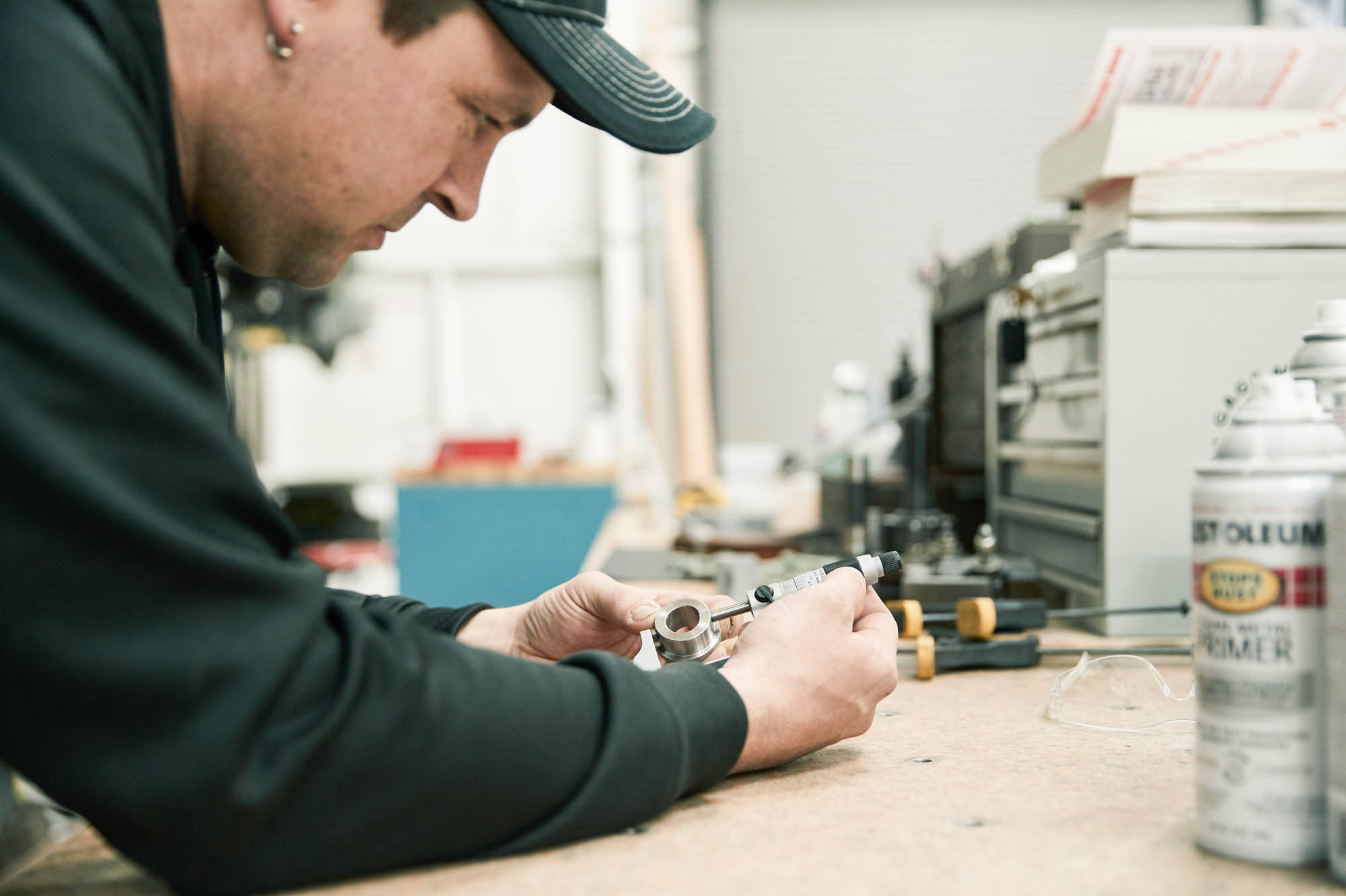 Boom Supersonic engineer wearing a baseball cap measuring a metal aircraft component at a workbench beside a can of Rust-Oleum primer