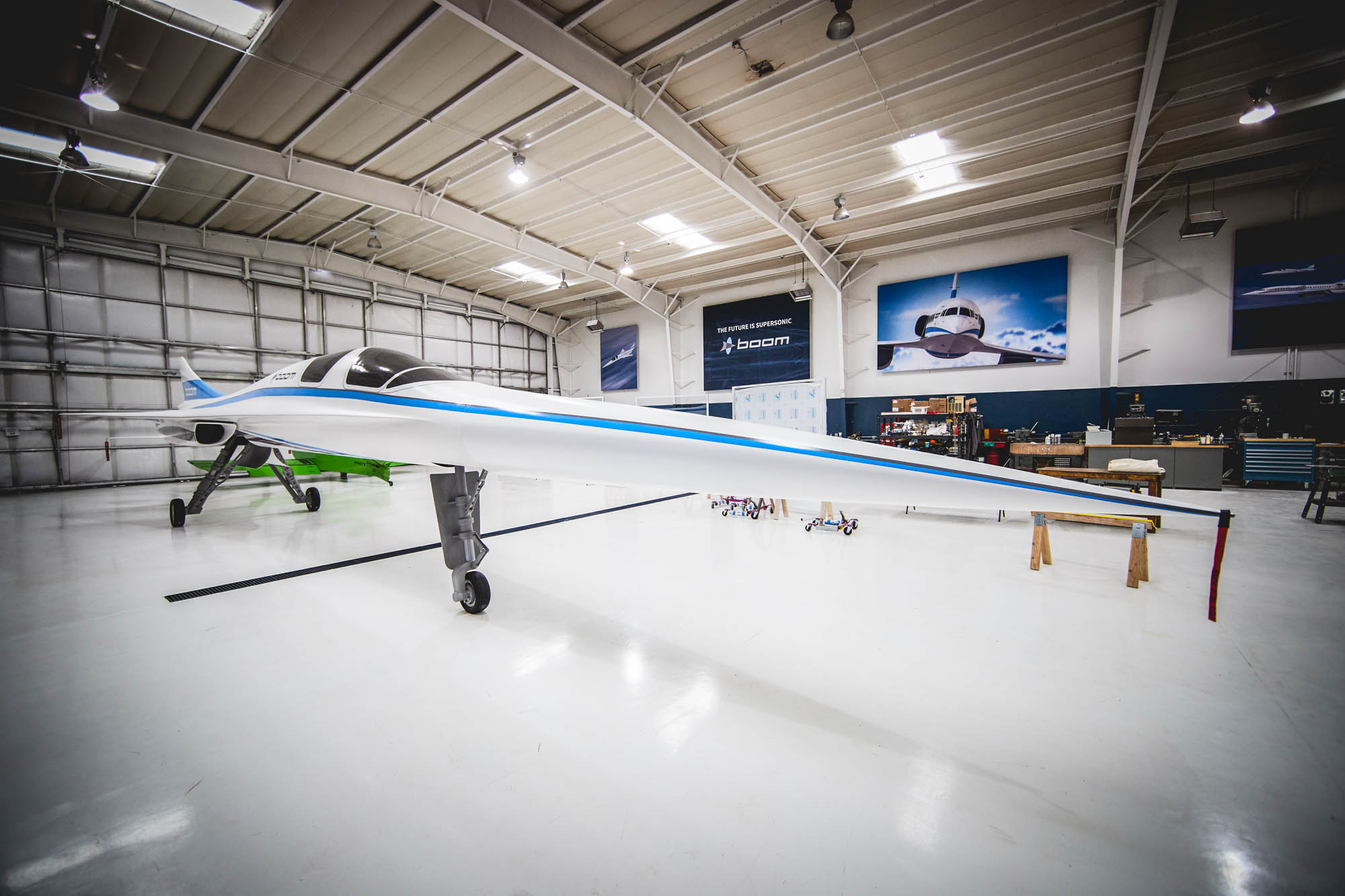 Side view of the white Boom XB-1 supersonic prototype aircraft with a long delta wing parked inside a hangar with Boom signage on the wall