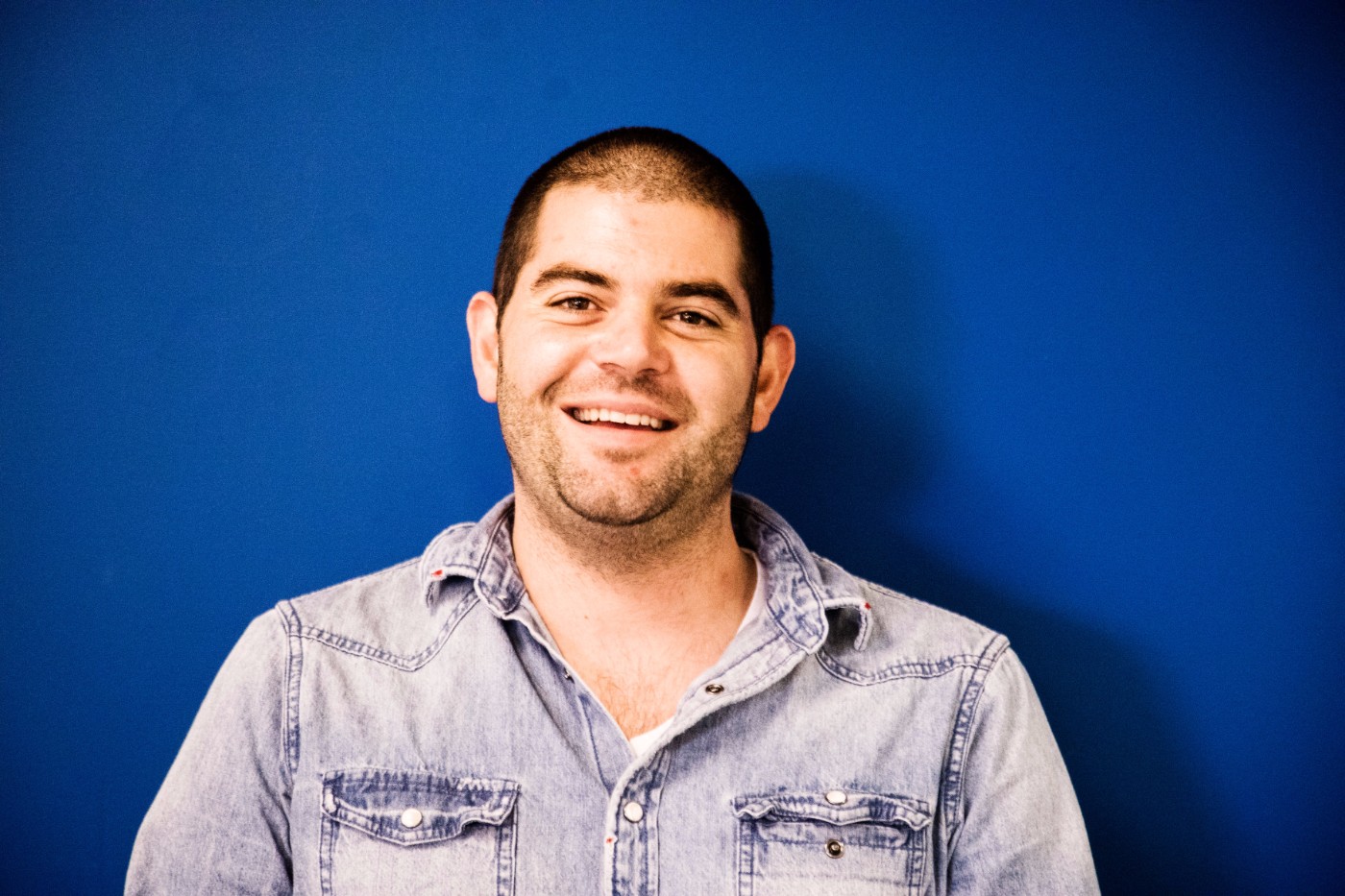 GoShare team member portrait - smiling man with short dark hair wearing denim shirt against blue backdrop