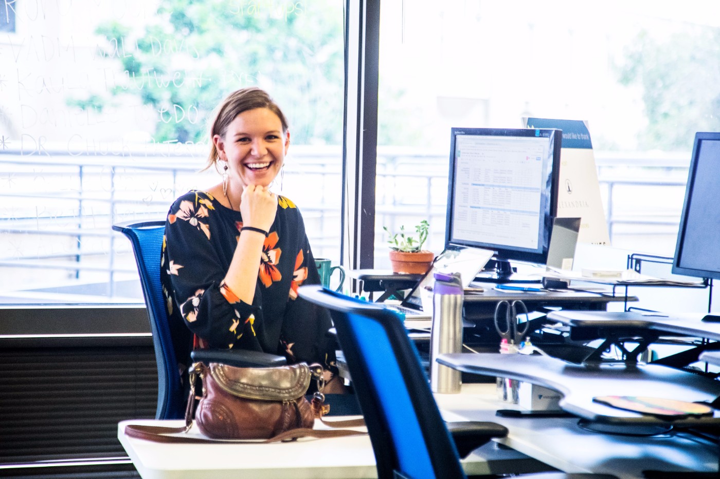 GoShare team member smiling at her desk in a brightly lit office with computer monitors and notes on window