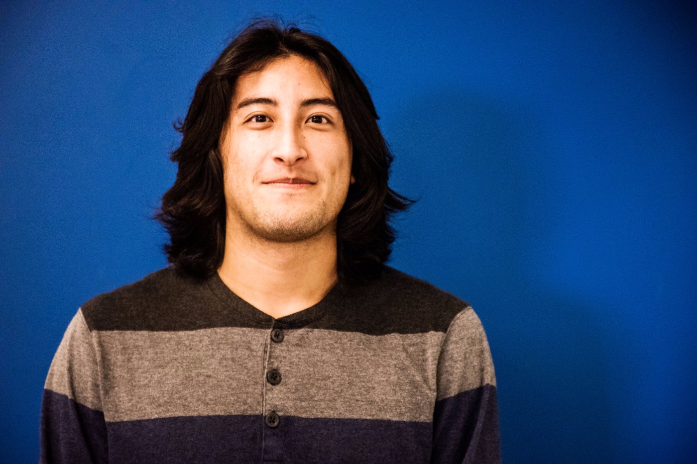 GoShare team member portrait - young man with long dark hair wearing striped henley against blue backdrop