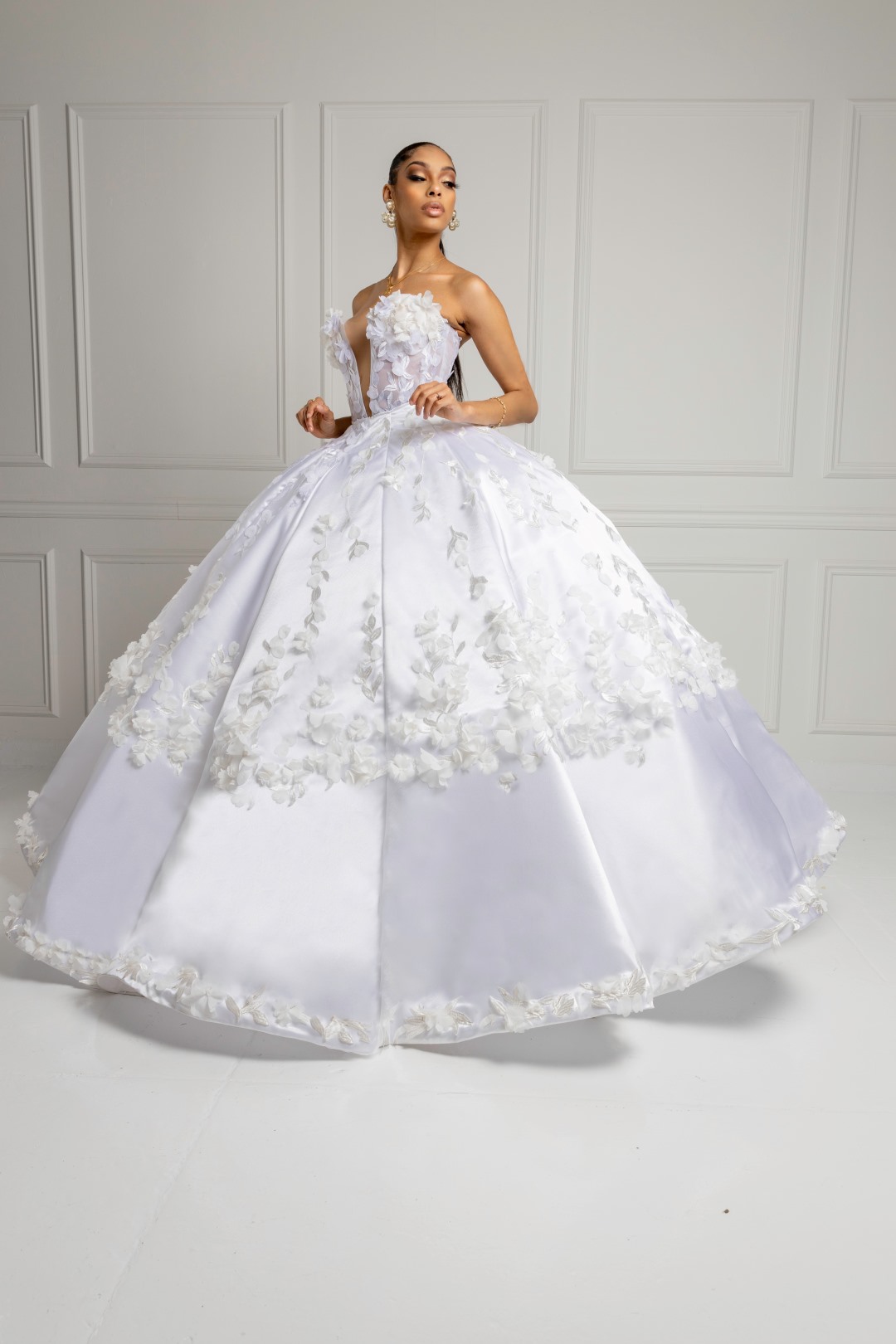 Woman in a white ball gown with floral appliqué standing against a paneled white wall.