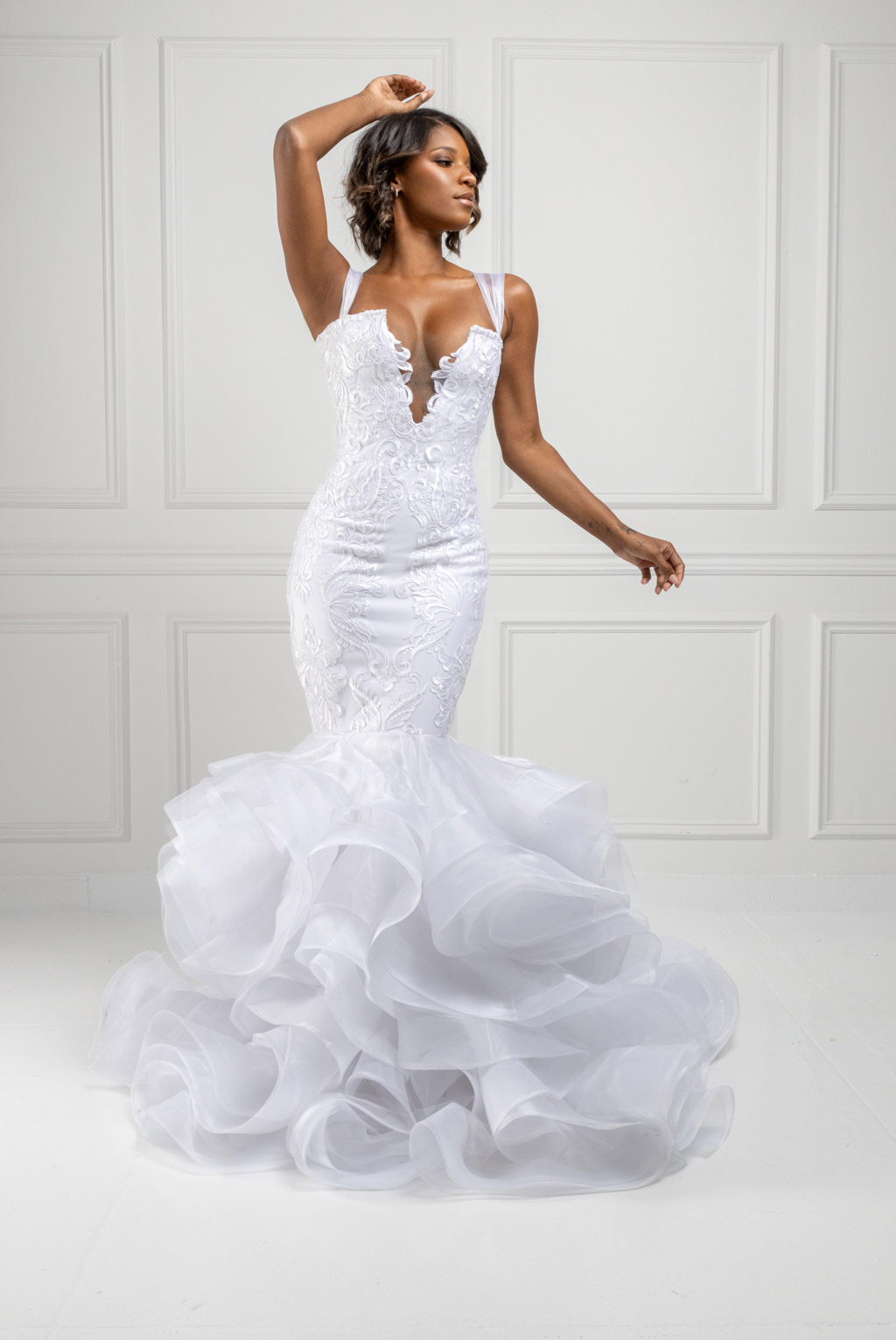 Woman in a white lace mermaid-style wedding dress with ruffled tulle skirt posing in front of white paneled wall.