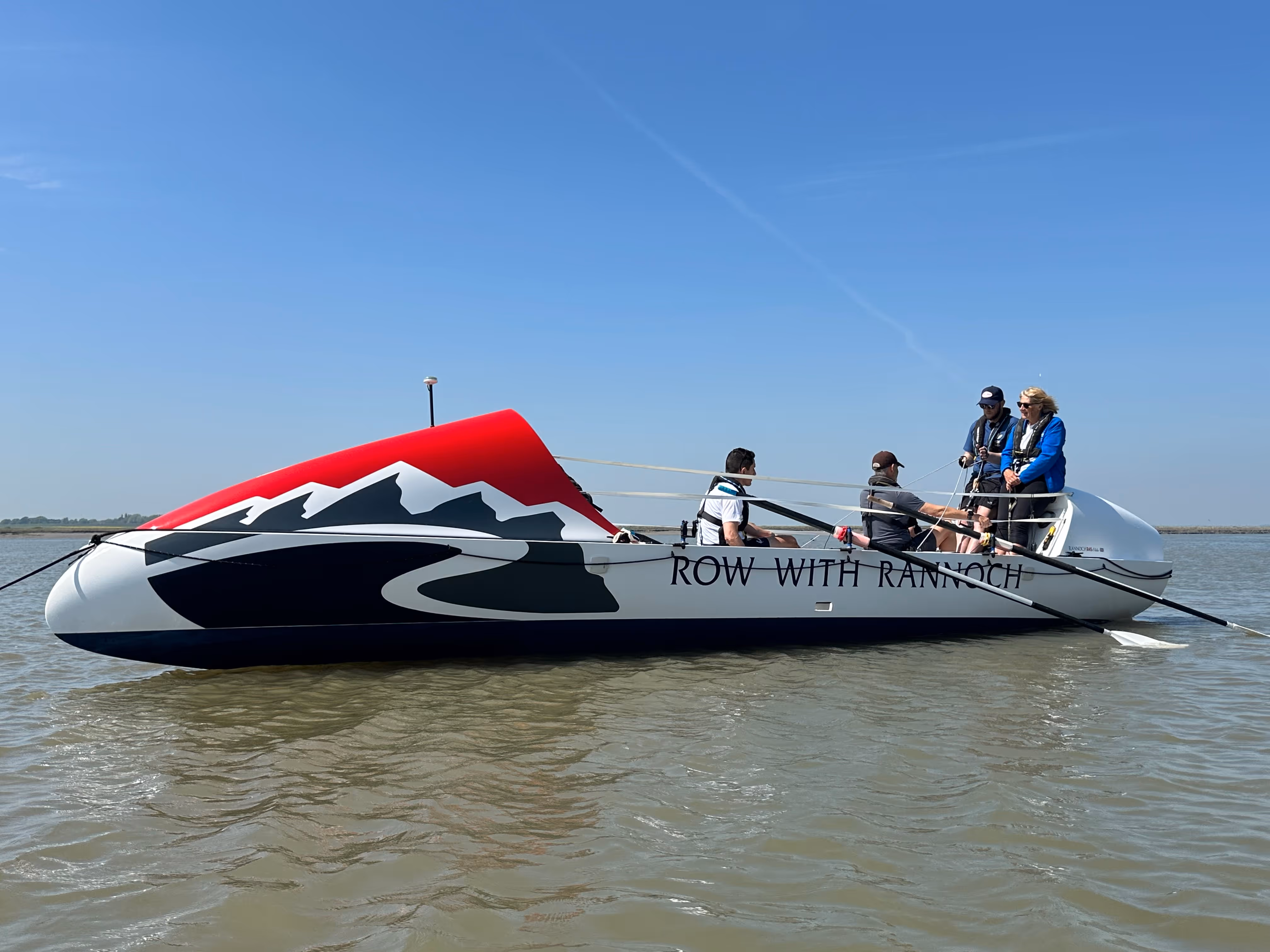 Four people sitting in a boat named "Row With Rannoch" on a calm body of water.