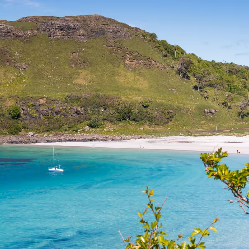 A sailboat sits on calm turquoise water near a green hillside and white sandy beach.