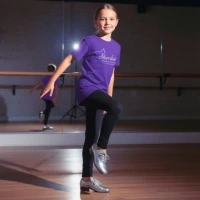 Young girl in purple shirt and black leggings practicing tap dance on one foot in a dance studio with mirrors.