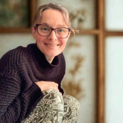 Smiling woman with glasses wearing a dark sweater and patterned leggings seated indoors.