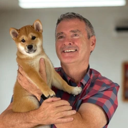 Smiling man in a red and blue plaid shirt holding a small Shiba Inu puppy indoors.