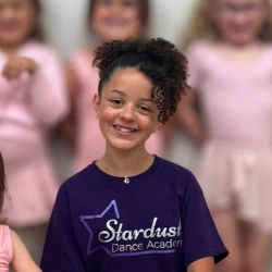 Smiling young girl with curly hair wearing a Stardust Dance Academy t-shirt, with blurred dancers in pink outfits behind her.