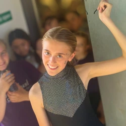 Smiling young girl in a sleeveless patterned top raising one arm indoors with blurred people in the background.