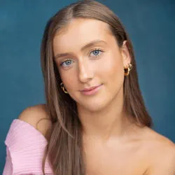 Young woman with long brown hair, blue eyes, and gold hoop earrings wearing a pink off-shoulder top against a blue background.