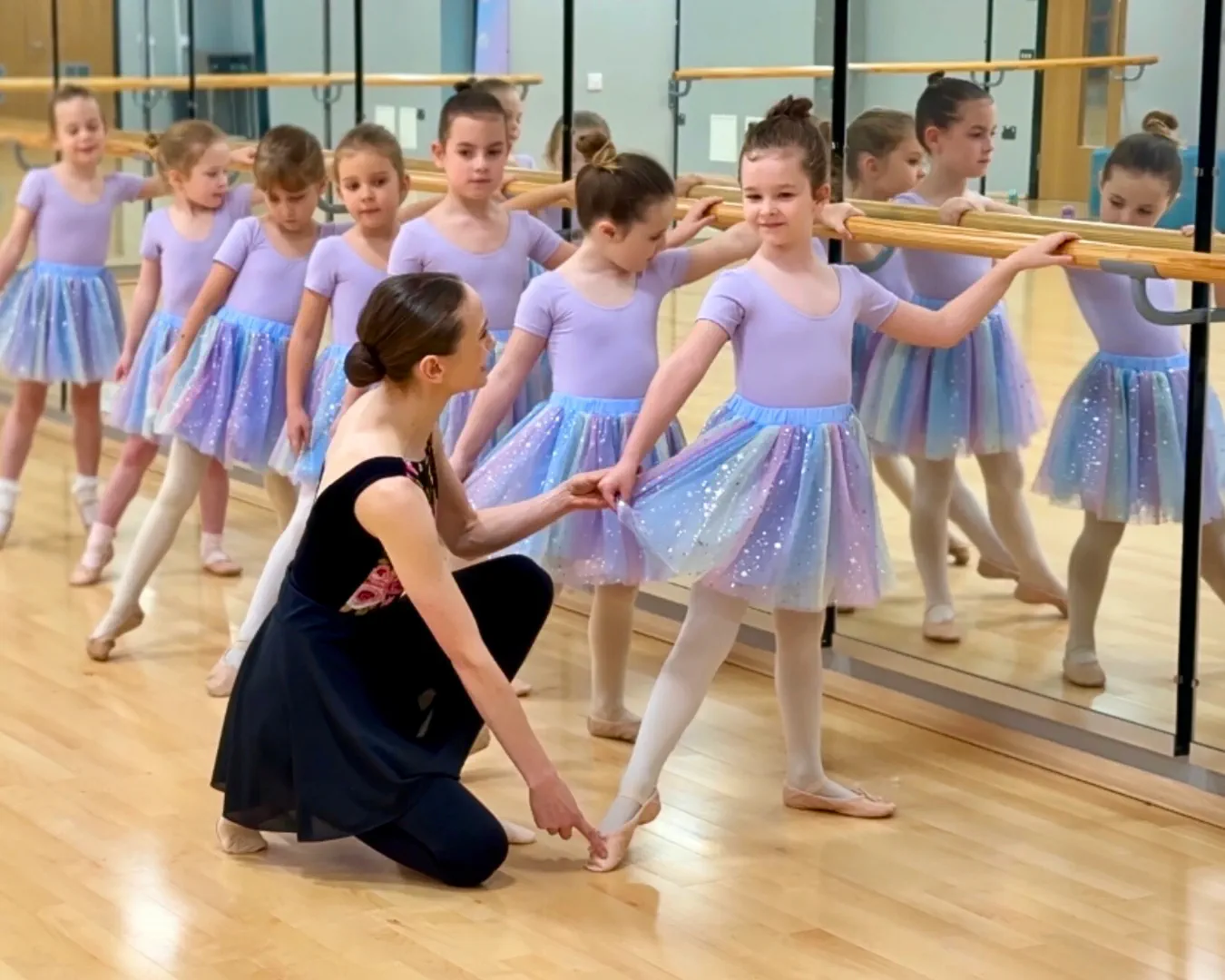 A primary ballet class student receiving support and guidance from ballet teacher whilst on the barre.