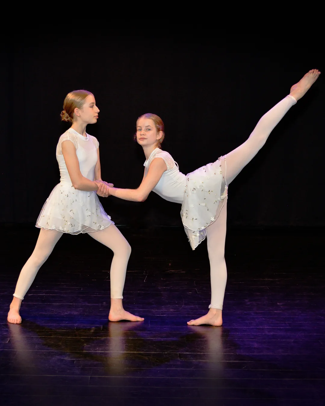Two young ballerinas in white costumes holding hands, one standing and the other balancing with one leg extended backward.