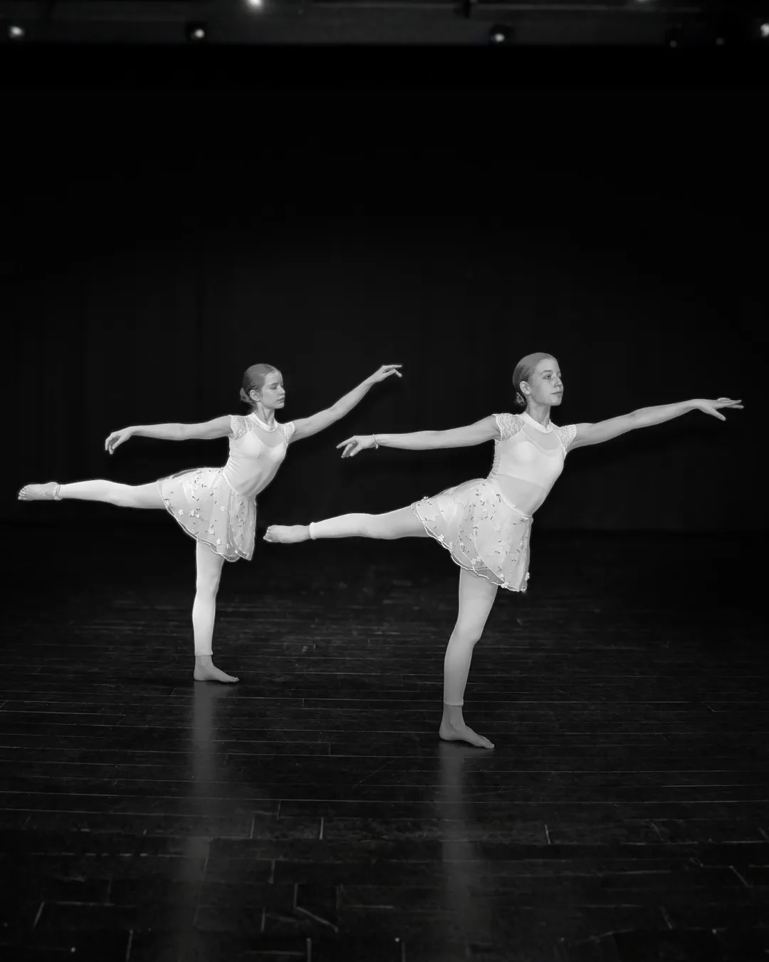 Two female ballet dancers in white costumes performing an arabesque pose on a dark stage.