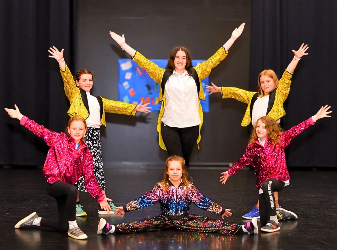 Six smiling children in colorful sequined costumes posing enthusiastically on stage during a musical theatre performance.
