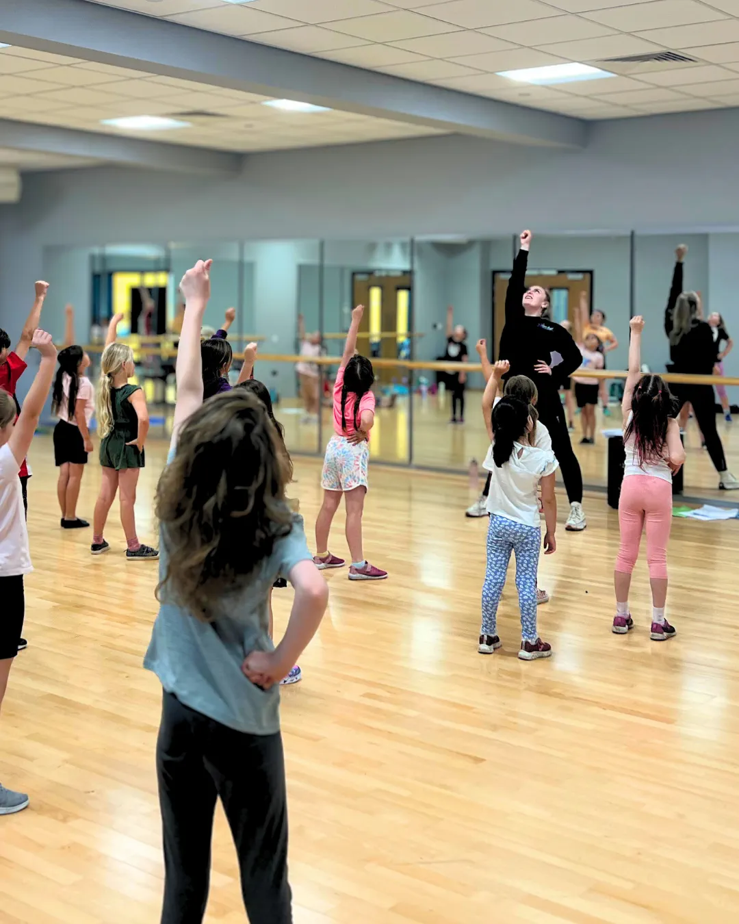Children in a dance class with arms raised, following an instructor in a mirrored studio.