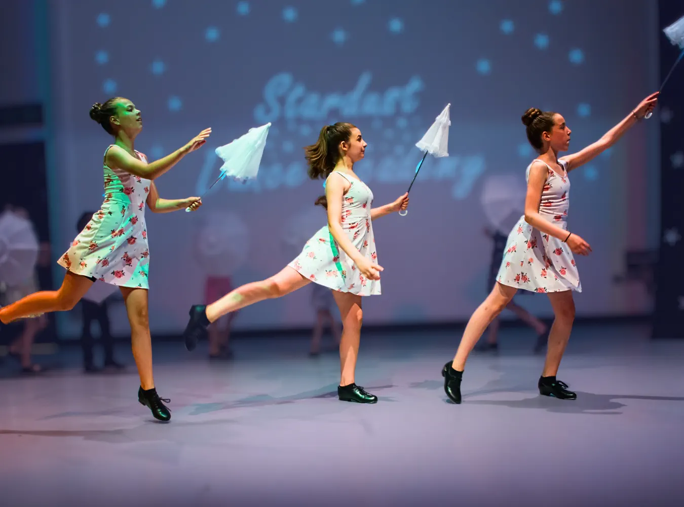 Three young female dancers in floral dresses performing with closed white umbrellas on stage.