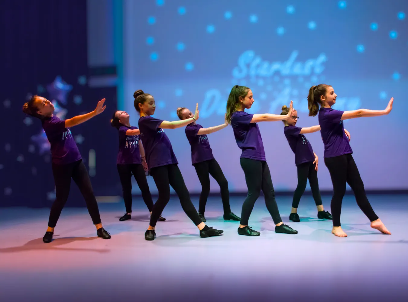 Group of young dancers in purple shirts performing a synchronized routine on stage with a blue background.