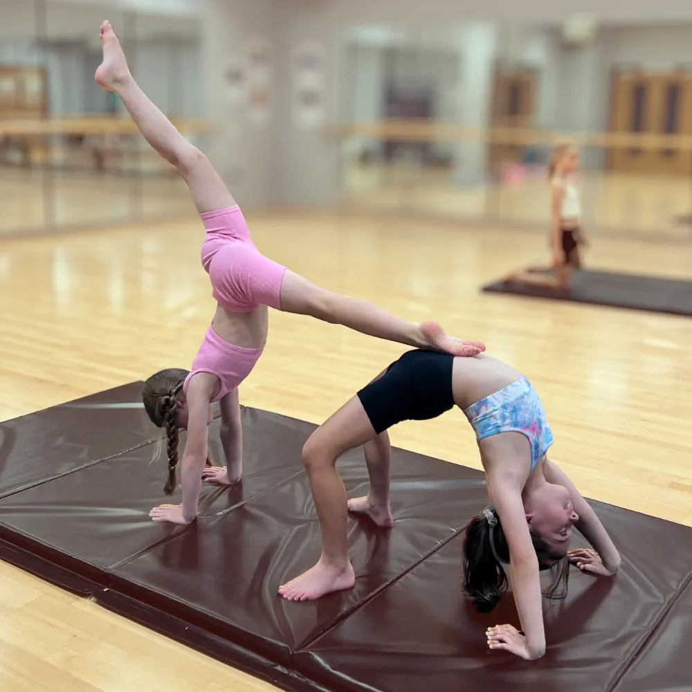 Two young girls practicing acro yoga on brown mats in a gym with mirrored walls and wooden floor.