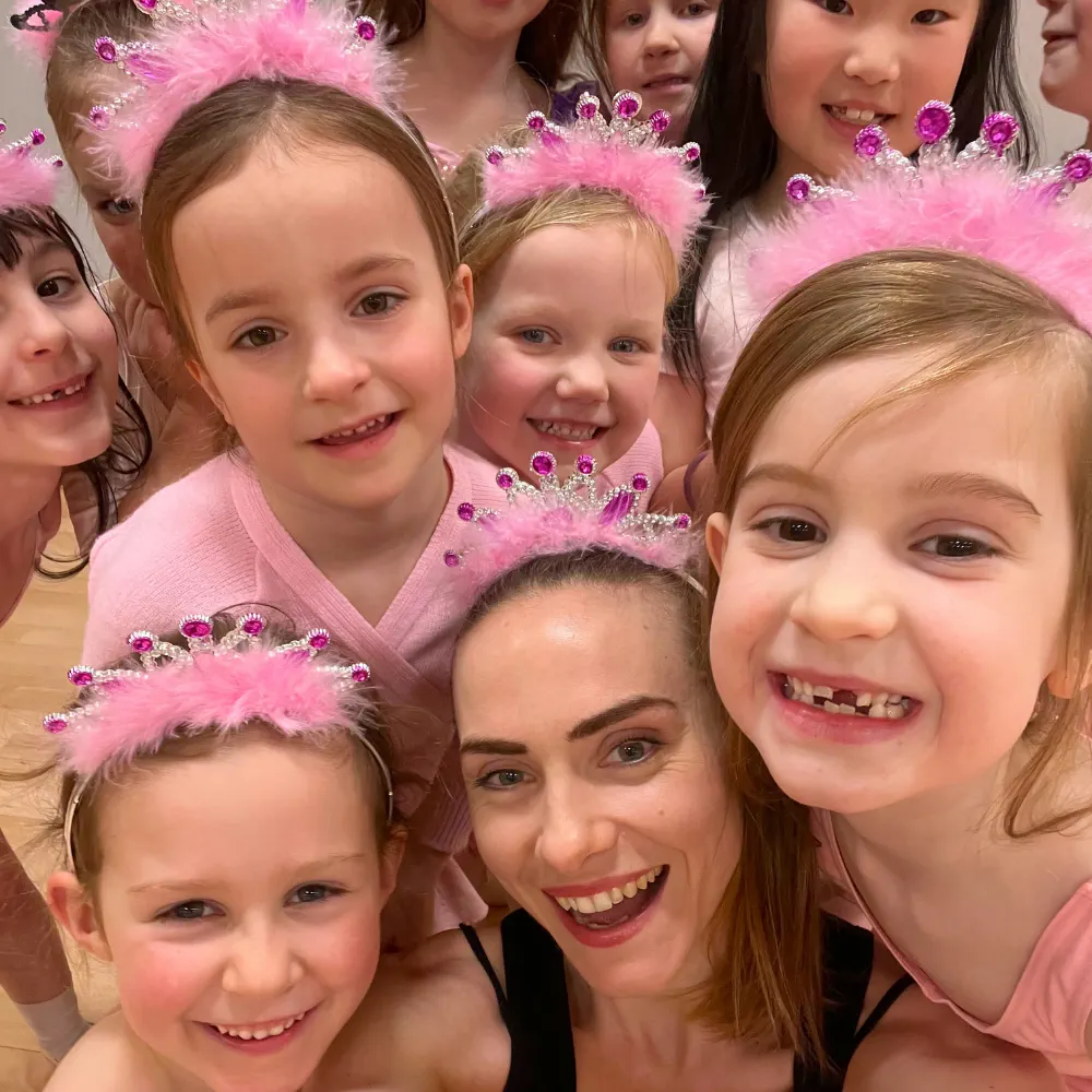 Group of smiling children and a woman wearing pink princess tiaras, posing closely together.