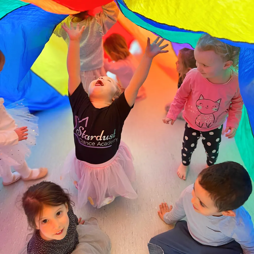 Group of young children playing under a colorful parachute indoors, one child joyfully reaching up.