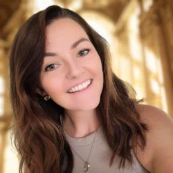 Smiling woman with long brown hair wearing a light gray top and necklace in a warmly lit indoor setting.
