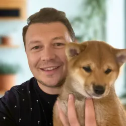 Smiling man holding a Shiba Inu puppy in a bright room.
