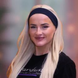 Smiling woman with long blonde hair wearing a black headband and black top, blurred background.