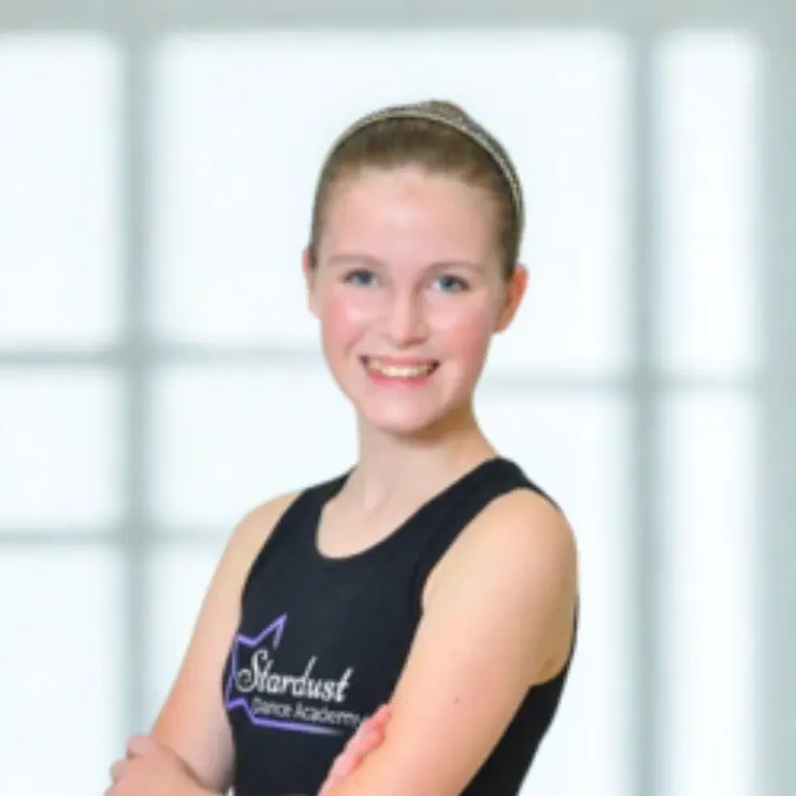 Smiling young girl with a headband and black tank top with Stardust Dance Academy logo, standing with arms crossed.
