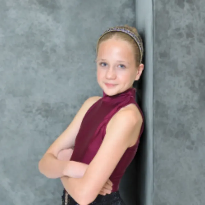 Young girl with a braided headband wearing a sleeveless maroon top, standing with arms crossed against a gray textured wall.