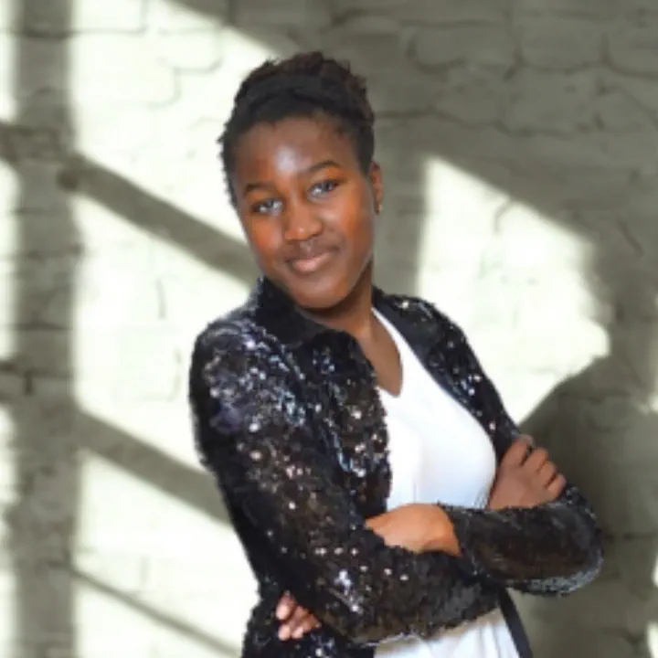 Smiling young woman with arms crossed wearing a black sequined jacket and white top, standing in front of a light brick wall with shadows.