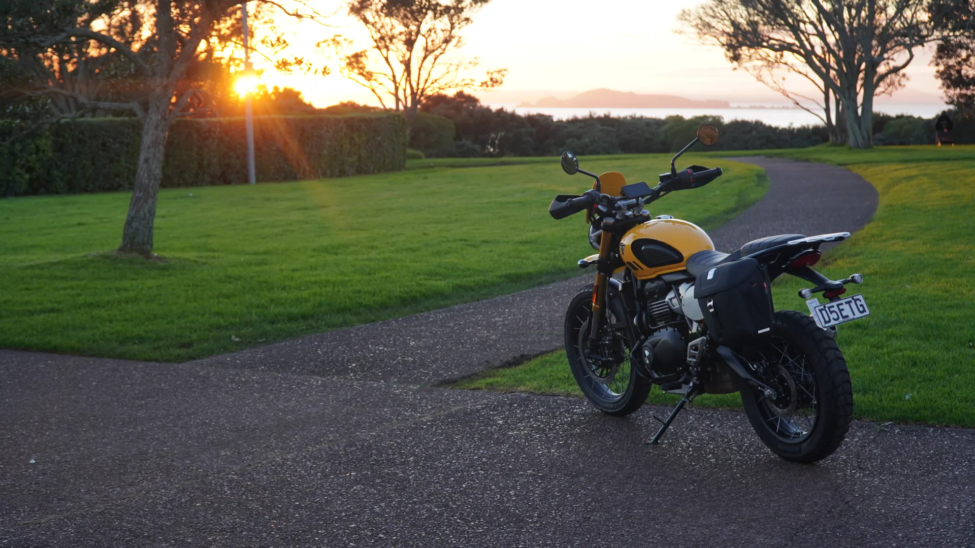 Yellow Triumph Scrambler 400XC parked on a coastal path at sunset, East Auckland