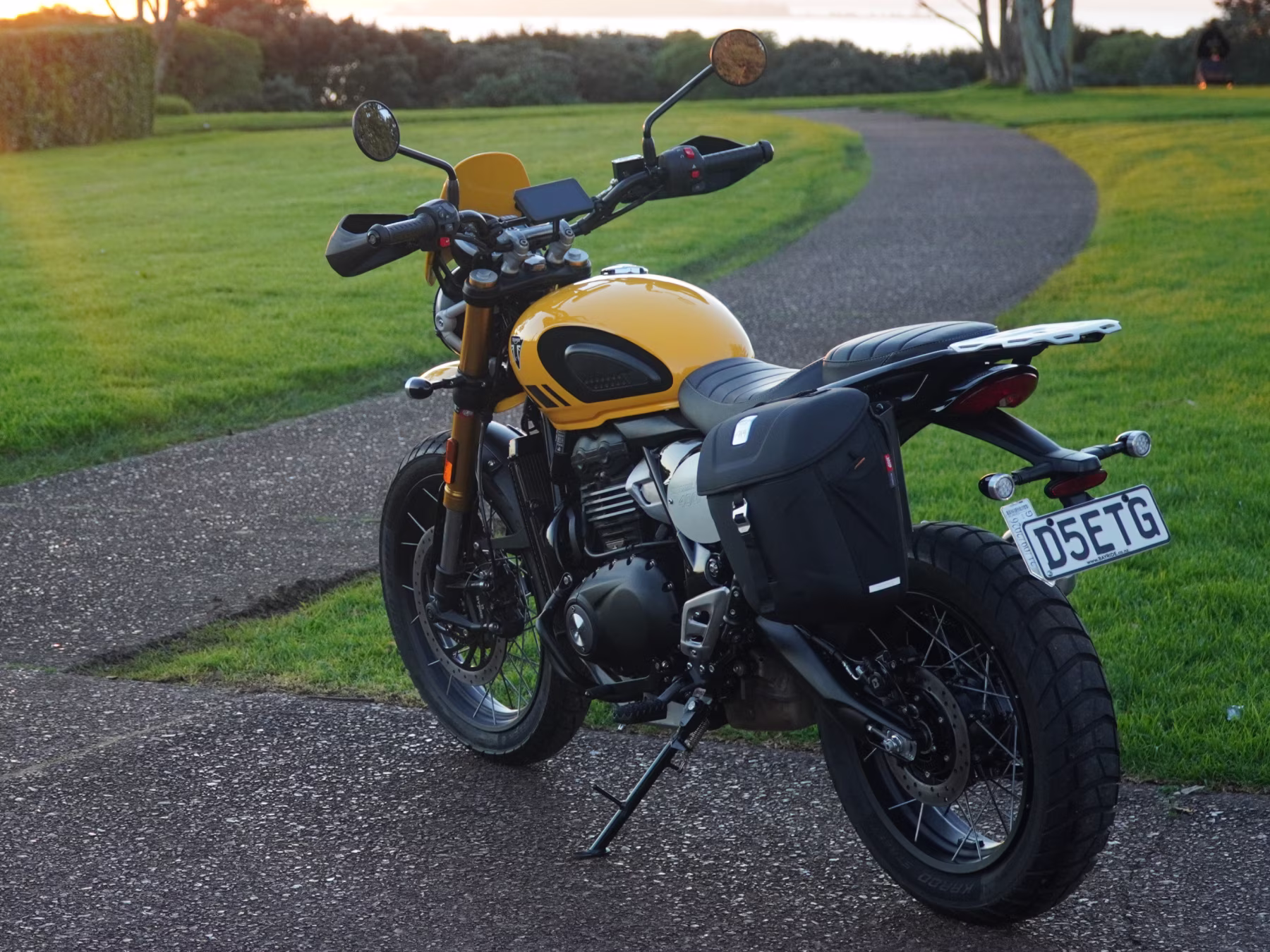 Yellow Triumph Scrambler 400XC with a black soft pannier, parked on a coastal path at sunset