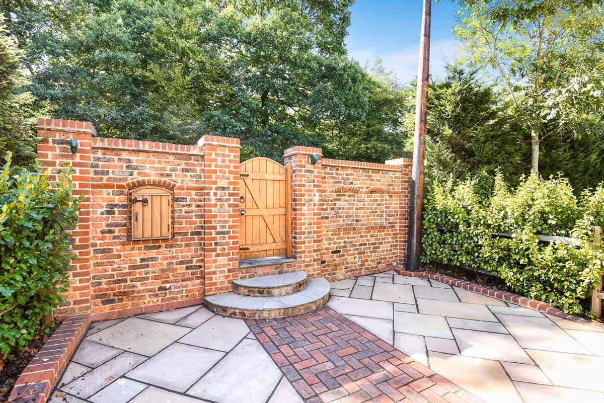 Brick wall with a wooden gate and steps, surrounded by trees and shrubs in a paved area.