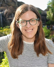 A woman with glasses and long brown hair smiles at the camera, with a blurred natural background.