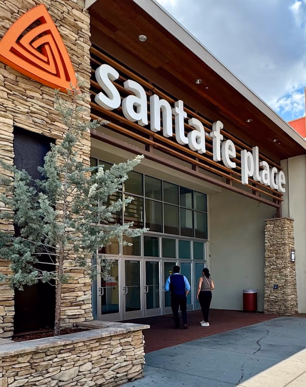 Entrance of Santa Fe Place shopping center with two people walking toward the glass doors and a tree in a stone planter.