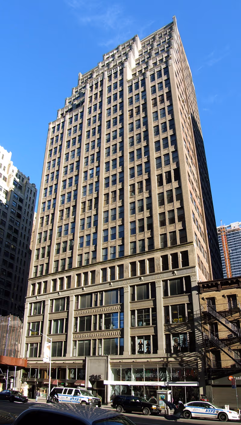 Tall multi-story commercial building with American Apparel store at street level and NYPD vehicles parked outside under clear blue sky.