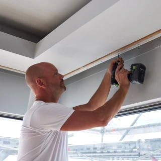 Man in white shirt using a cordless drill to install a concealed blind system on ceiling.