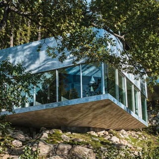 Modern glass house with concealed blinds surrounded by trees and rock landscaping.