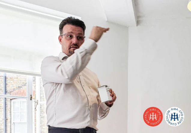 Man in glasses demonstrates a small white concealed blind system part inside a bright room.