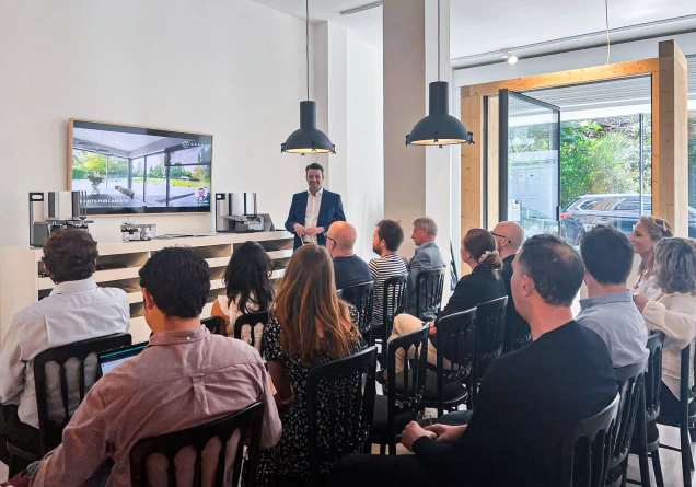 Man giving a presentation to an audience seated in a modern room with a TV screen and large windows.