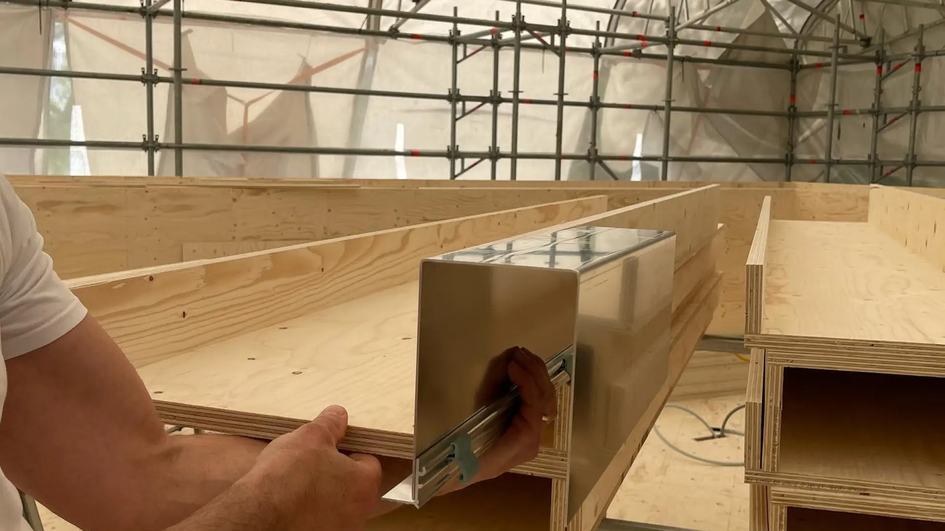 Close-up of hands installing a metal blind head box on a timber frame under construction.