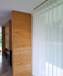 Sheer white vertical blinds partially covering a floor-to-ceiling window beside wooden wall panels.
