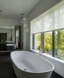 Modern bathroom with white freestanding tub under a large window with a roller blind and pendant lights.