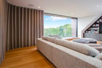 Modern living room with large gray sectional sofa, brown curtains, and floor-to-ceiling window showing greenery.
