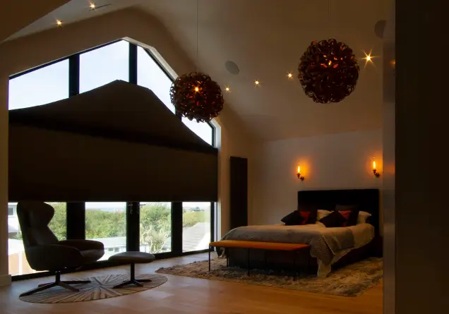 Bedroom with vaulted ceiling, angled windows with blackout blinds, hanging lights, bed, and lounge chair.