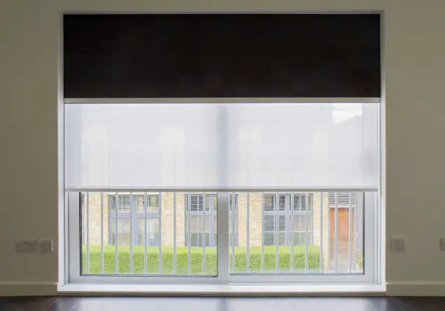 Window with double roller blinds: black blackout on top and white screen below showing a balcony railing.