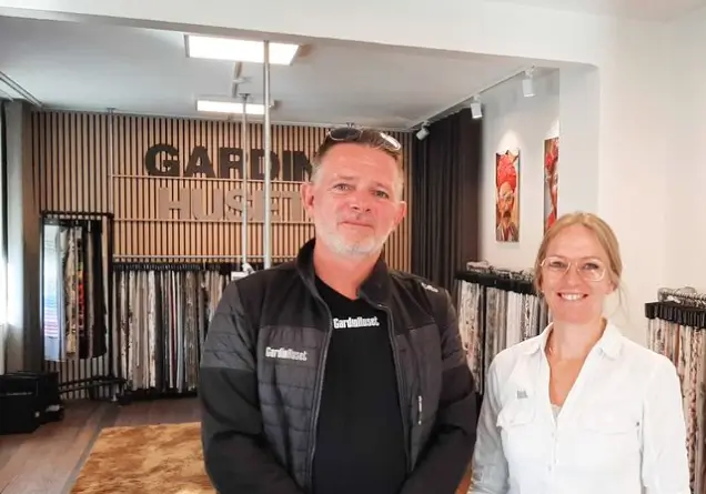 Two smiling adults standing in a fabric showroom with patterned textile samples behind them.