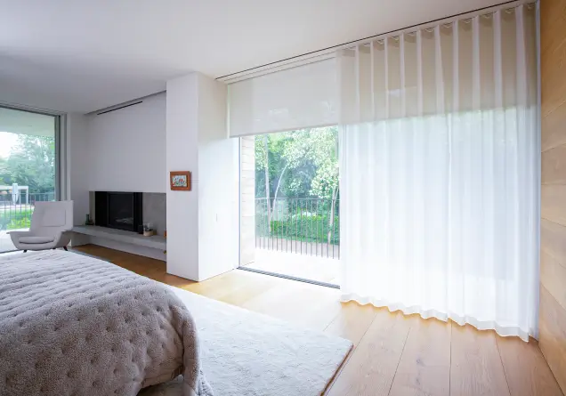 Bright bedroom with floor-to-ceiling window covered by sheer wave curtains and light wooden floor.
