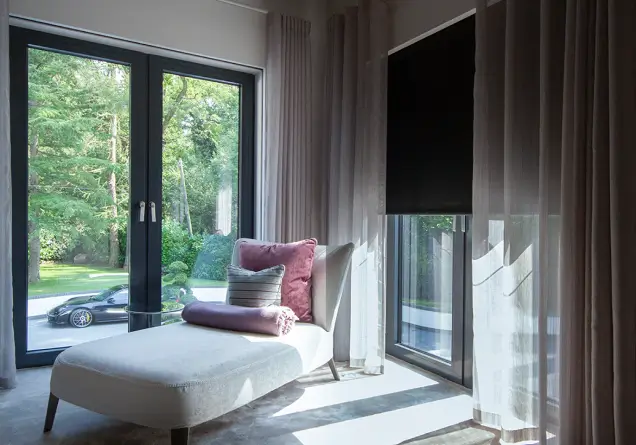 Modern room with beige chaise lounge, velvet cushions, sheer curtains, and black motorized window blind.